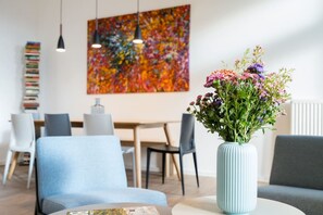 Una sala de estar moderna con un sofá azul, una mesa de centro redonda y un jarrón de flores. Se observa un comedor con una mesa y sillas al fondo.
