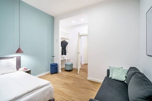 Ein modernes Hotelzimmer mit einem Bett, einem grauen Sofa, einem kleinen Tisch und einem blauen Koffer.