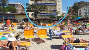En strand med solstolar och parasoller, en byggnad med balkonger och en klarblå himmel.
