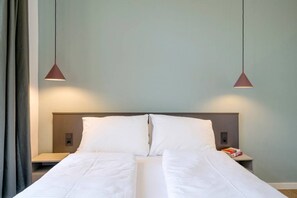 En pænt redt seng med hvide sengetæpper, to sengeborde med lamper og en lyseblå væg.