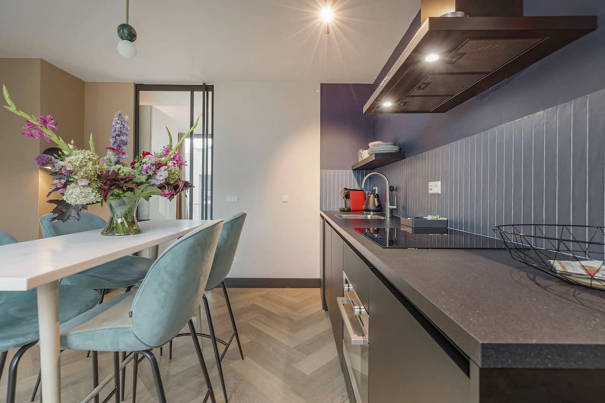 Una cucina moderna con zona pranzo, caratterizzata da un pavimento in parquet a spina di pesce, un'isola centrale con lavello e un vaso di fiori sul tavolo.
