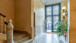 Un escalier menant à une porte surmontée d’un portail en fer forgé, avec une colonne en marbre et un pot de fleurs.