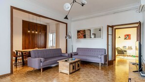 Un salon comprenant un canapé d’angle, une table basse en bois, un coin repas avec une table et des chaises, et un coin cuisine avec des placards et un plan de travail.