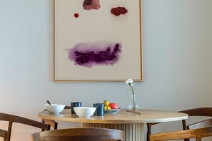 A dining table with a bowl, mug, and fruit, and a framed abstract painting on the wall.