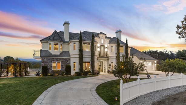 A large, two-story house with a stone facade, multiple chimneys, and a curved driveway leading to a gated entrance. - Chateau Vino - French Style Chalet (Temecula)