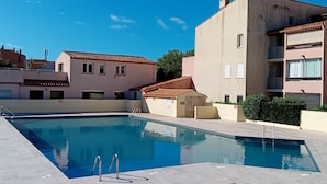 Una piscina con un borde empedrado, rodeada de edificios con balcones y un cielo azul despejado.