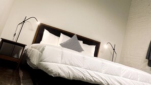 Un lit bien fait, avec une literie blanche et un coussin à motifs noir et blanc, dans une pièce aux murs blancs et dotée d’une tête de lit en bois.