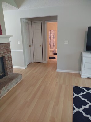 A spacious living room with hardwood flooring, a fireplace, a television on a white cabinet, and a door leading to a bathroom with a shower curtain.