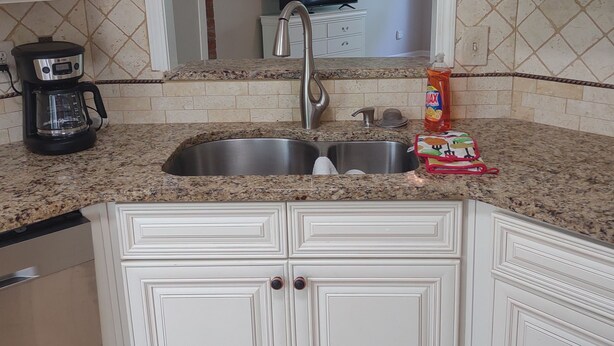 Una cocina con gabinetes blancos, encimera de granito, pileta de acero inoxidable, cafetera y televisión al fondo.