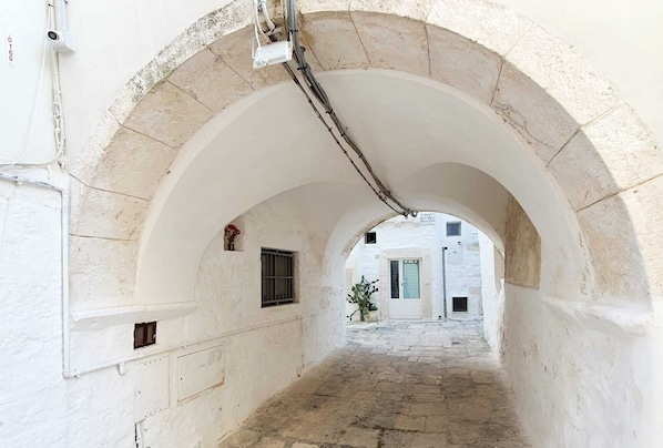 A narrow, arched corridor with stone walls and a tiled floor, leading to a building with a door and windows.