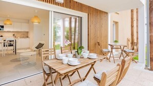 Un comedor moderno con mesas y sillas de madera, una cocina al fondo y un amplio ventanal corredero que da a un espacio exterior.