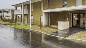 Un edificio residencial moderno con techo plano, vestíbulo cubierto y un pequeño jardín delimitado por una valla de madera.