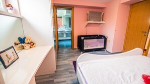 Un dormitorio con cama, escritorio con un osito de peluche, televisor y baño visible a través de una puerta abierta.