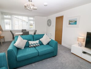 A teal sofa with white and patterned cushions, a dining table with chairs, a wooden door, a clock, a framed picture, and a TV on a white cabinet. - Bryn Derw Bach (Moelfre)