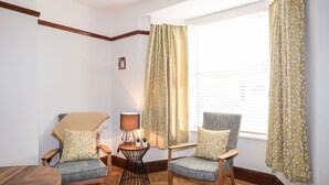 Um quarto acolhedor com duas cadeiras de madeira, uma mesa redonda pequena com uma luminária e uma janela com cortinas.