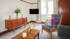 Uma sala de estar com um armário de madeira, uma televisão na parede, uma cadeira com almofada e um vaso com flores sobre uma mesa.