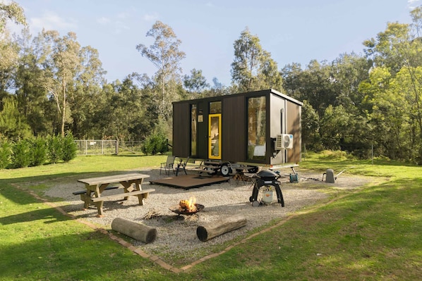 A compact, modern mobile home with a flat roof, a deck, and a fire pit in a grassy area.
