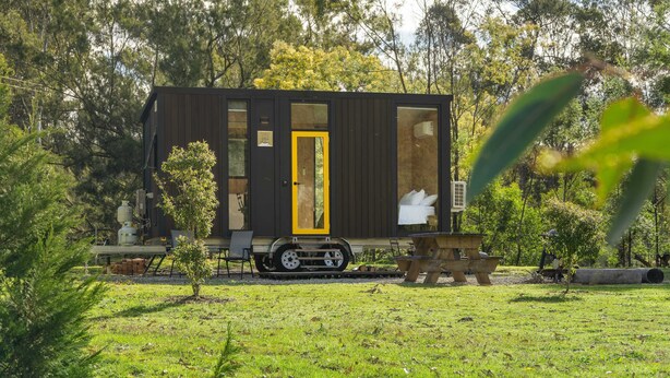 Een compacte, donkerkleurige stacaravan met een gele deur, gelegen in een groene omgeving met bomen op de achtergrond.