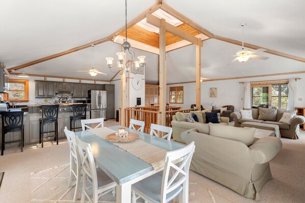 A spacious living area with a dining table, sofas, and a kitchen area with wooden beams and a chandelier. - Moby Duck: Ocean and Sound Views | Pet-Friendly (Kitty Hawk)