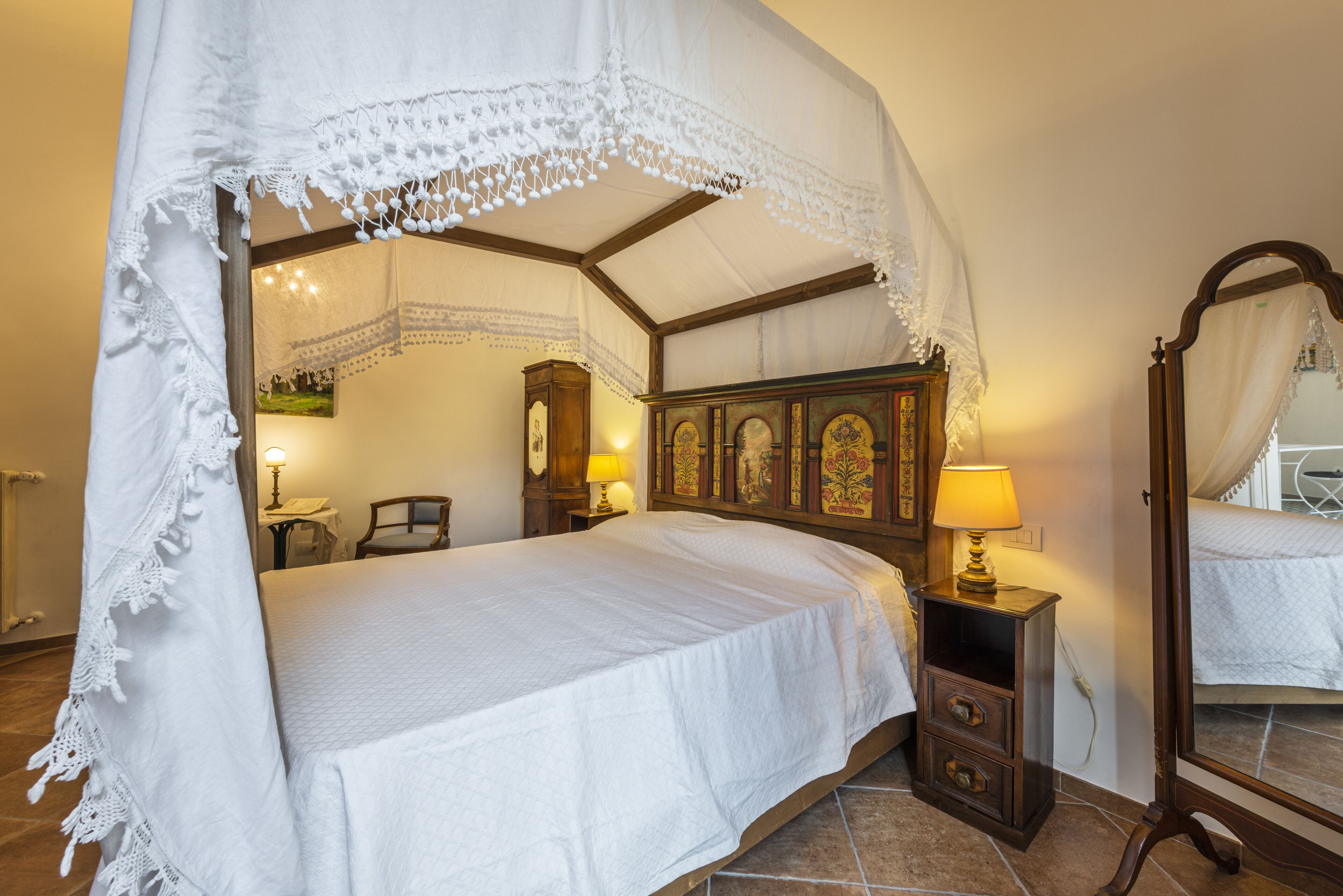 Quarto com cama de dossel, cabeceira de madeira, mesa de cabeceira com abajur e espelho.