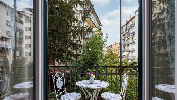 En balkon med to runde borde og stole, en plantekasse og udsigt til bygninger og træer.