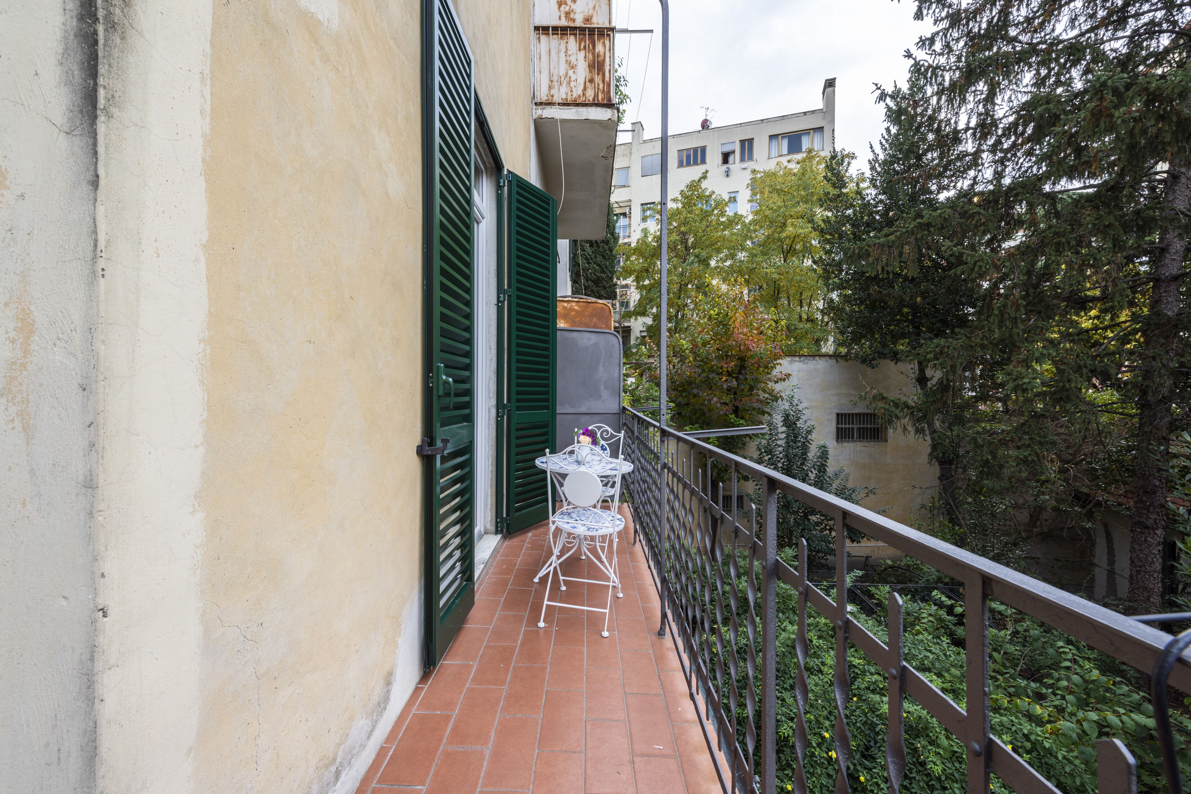 A balcony with a table and chairs, a green shutter door, and a view of trees and buildings.