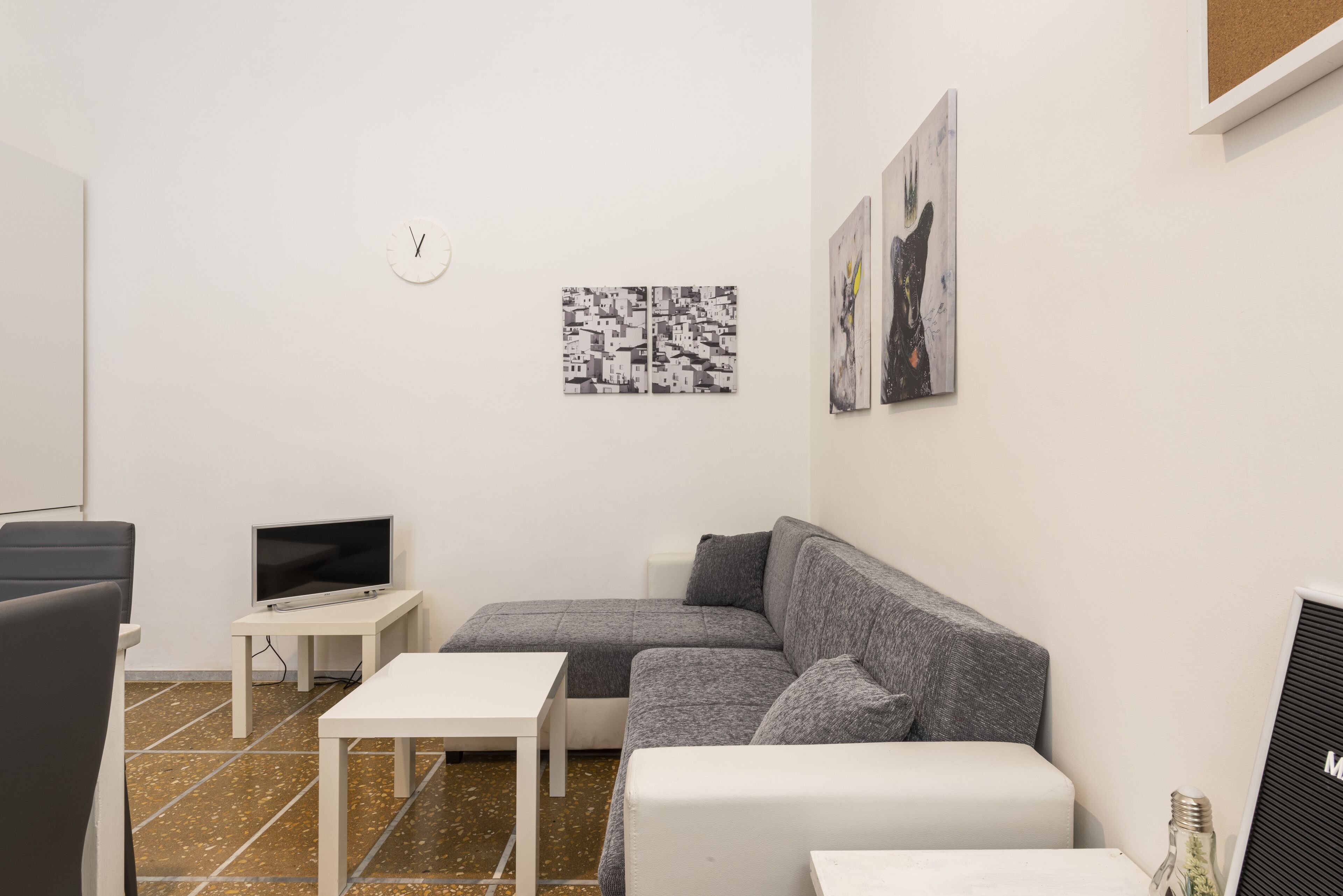 A modern living room with a grey sectional sofa, a white coffee table, a flat-screen TV on a small table, and a wall clock.