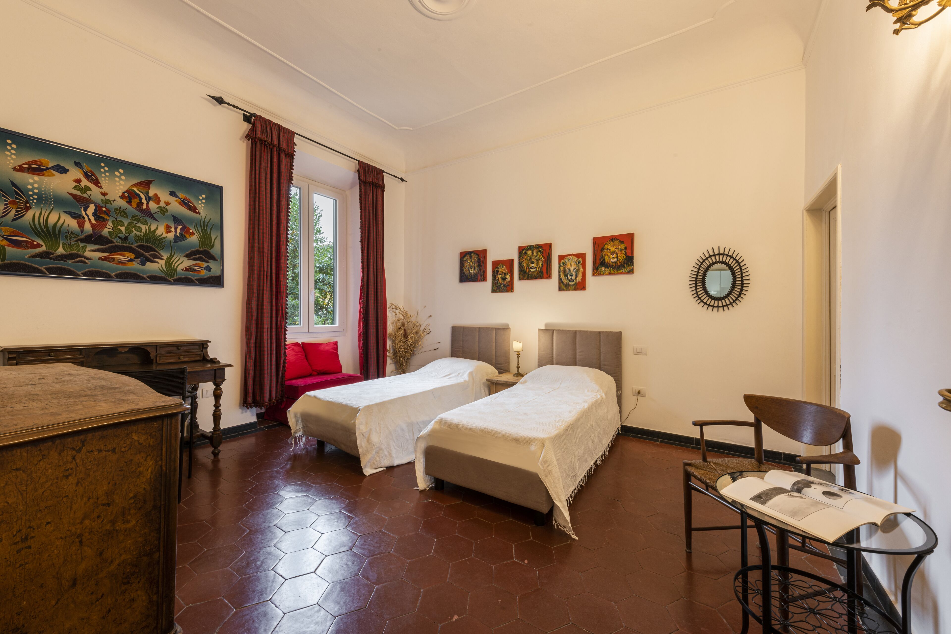 A room with two beds, a desk, and a chair. There is a painting on the wall and a window with curtains.