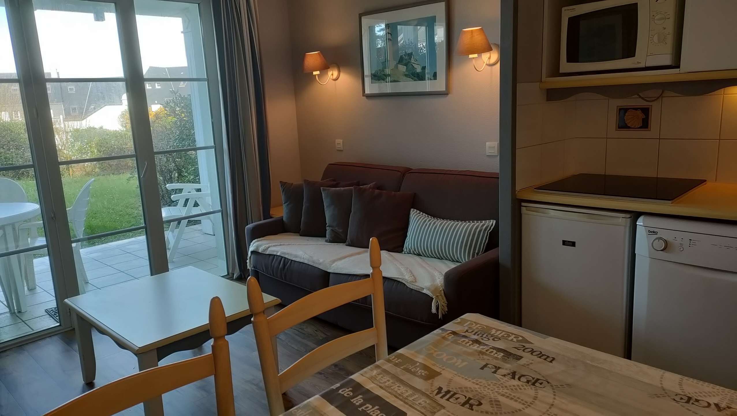 A compact living space with a dining table, a sofa, a small kitchen area with a microwave and a refrigerator, and a view through a glass door to an outdoor patio.