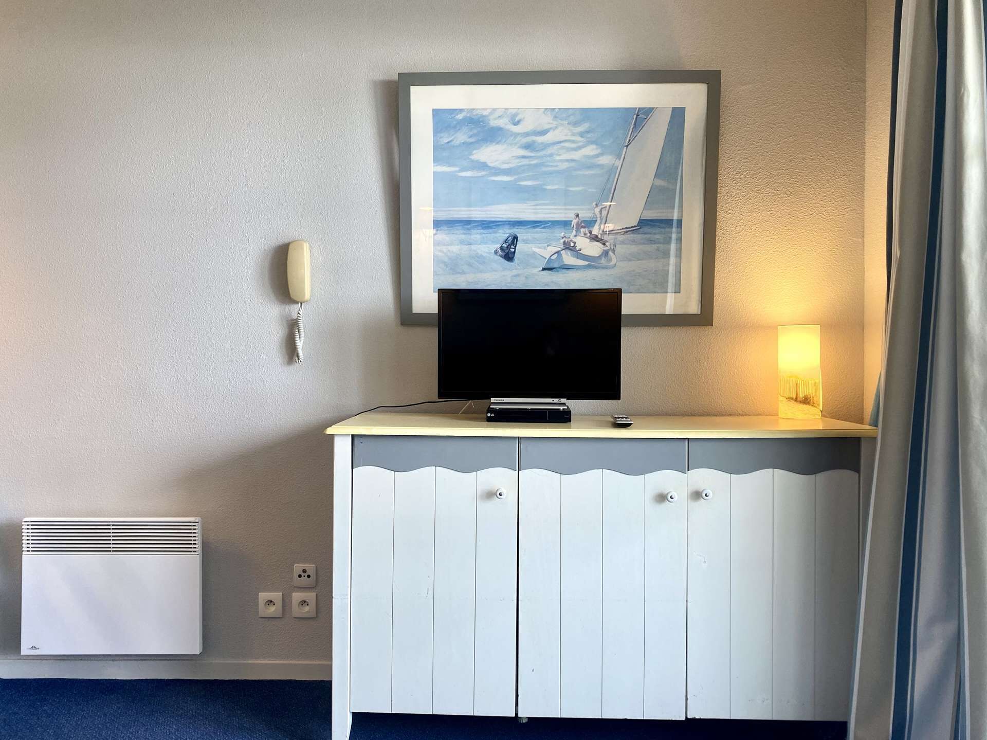 Une chambre d’hôtel avec un téléviseur posé sur un meuble blanc, un tableau encadré représentant un voilier, une lampe de chevet et un téléphone fixé au mur.