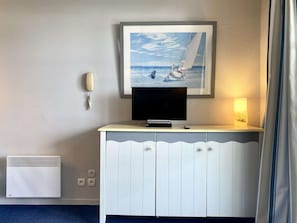 Quarto de hotel com televisão em um armário branco, um quadro com um veleiro, luminária de cabeceira e telefone fixo na parede.