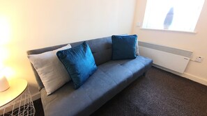 Ein graues Sofa mit zwei blauen Kissen, ein kleiner Tisch mit einer Lampe und ein Heizkörper in einem Raum mit einer schlichten weißen Wand und einem Fenster.