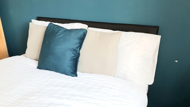 A bed with a white quilted cover, a dark headboard, and two pillows, one blue and one white. - Salford Ark Comfort Stays Near Trafford Centre (Manchester)