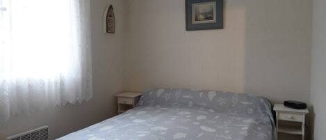 A bedroom with a bed, a nightstand, a small shelf, a window with sheer curtains, and a framed picture on the wall.