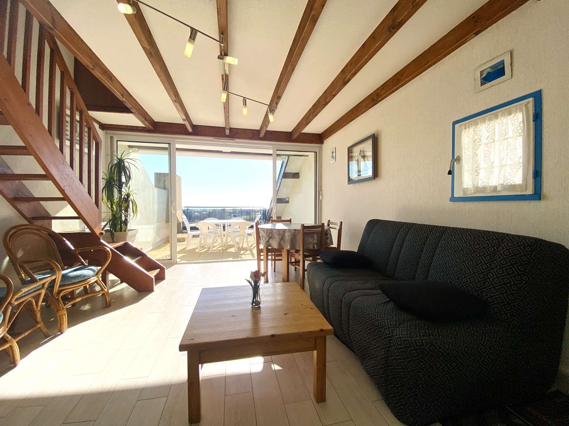 A spacious living area with a wooden staircase, a sofa, a dining table, and a balcony with outdoor seating.