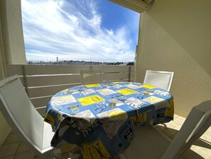 Una mesa redonda con mantel de tela azul y amarillo, dos sillas blancas y vista a un horizonte urbano a través de una ventana.