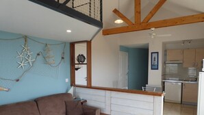 A cozy living area with a brown sofa, a patterned throw, and a wooden bench. A kitchen with white cabinets and a dining area is visible in the background. - Apartment for 4 sea view near Crouesty port (ARZON)