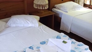Ein Hotelzimmer mit zwei Betten, beide mit weißen Laken und einer blau gemusterten Decke. Das hölzerne Kopfteil weist eine herzförmige Aussparung auf, daneben ein kleiner Nachttisch mit einer gehäkelten Lampe und ein Fenster mit Vorhängen.