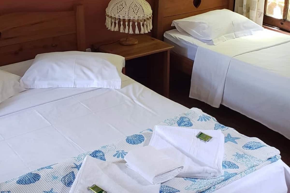 A hotel room with two beds, each with white linens and a blue patterned blanket. There is a wooden headboard with a heart-shaped cutout, a small wooden nightstand with a crocheted lamp, and a window with curtains.
