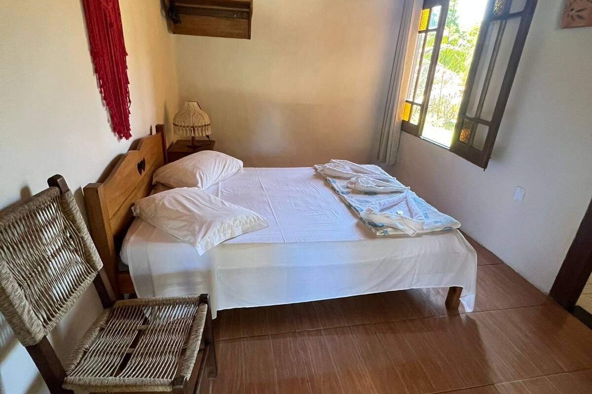 A bedroom with a wooden bed, a chair, a wall-mounted air conditioner, and a window with a view of greenery.