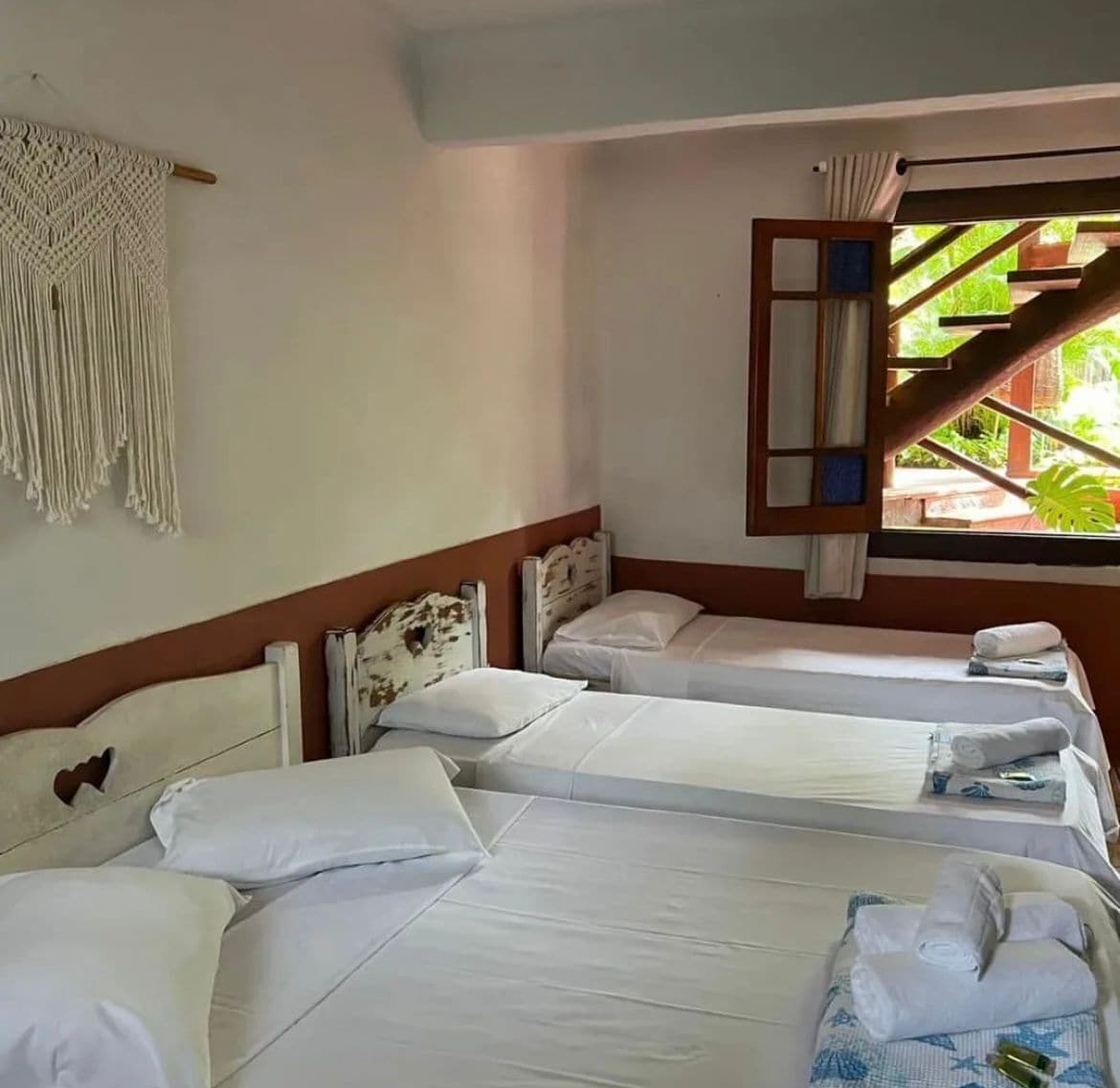 A hotel room with three single beds, a macrame wall hanging, and a window with a view of greenery.