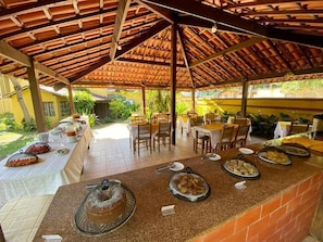 An outdoor dining area with a covered patio, tables set for meals, and a variety of dishes displayed on the counter. - Pousada Refugium (Itacaré)