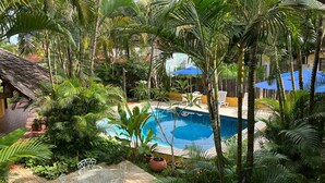 Ein Pool, umgeben von üppiger Vegetation und Palmen, mit einer gefliesten Terrasse und Gartenmöbeln.