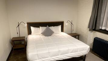 Een netjes opgemaakt bed met een witte deken, twee nachtkastjes met lampen, een houten hoofdbord, een radiator en een witte bakstenen muur.