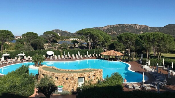 Una piscina grande y curvada con sillas de descanso y sombrillas, rodeada de árboles y montañas al fondo.