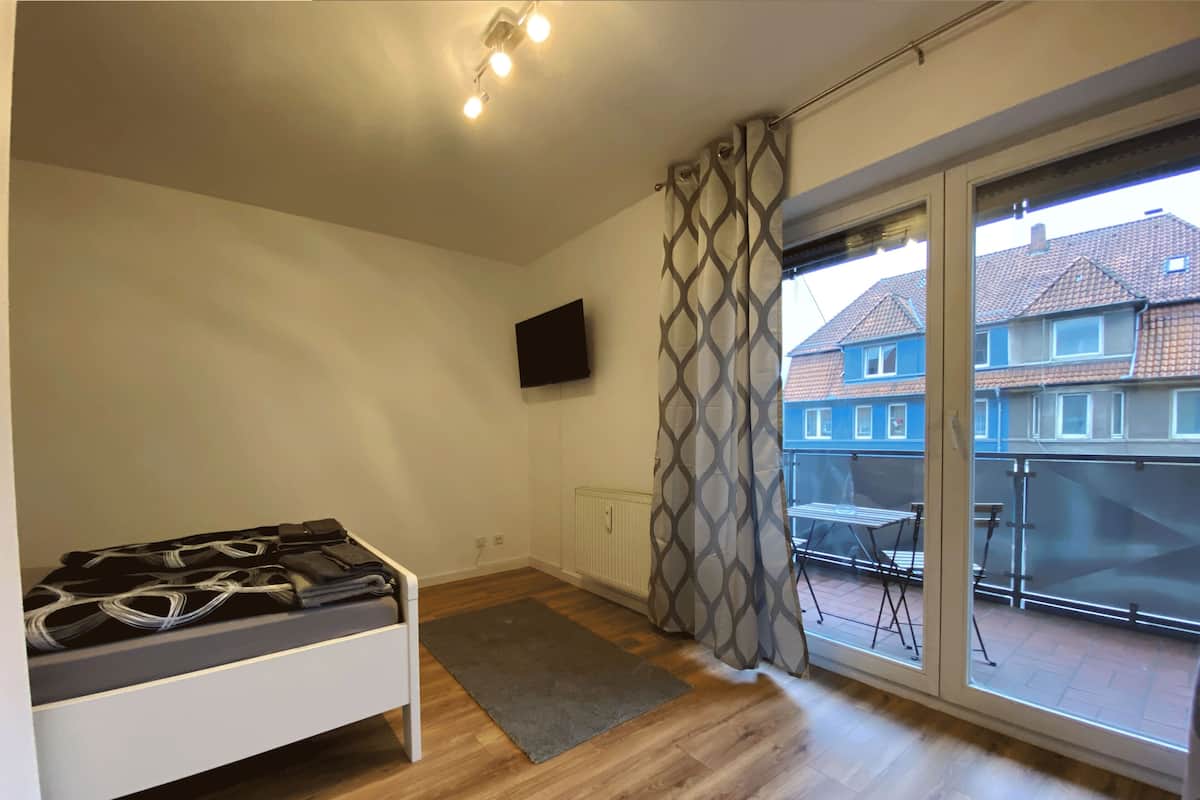 A bedroom with a bed, a TV, a window with a view of houses, and a balcony with a table and chairs.