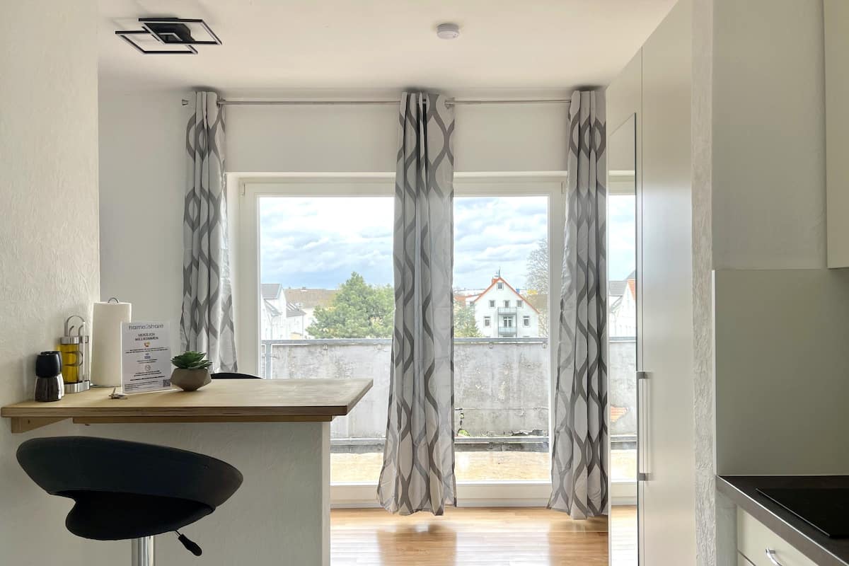 A modern kitchen with a wooden countertop, a bar stool, and a view of the outside through large windows with patterned curtains.