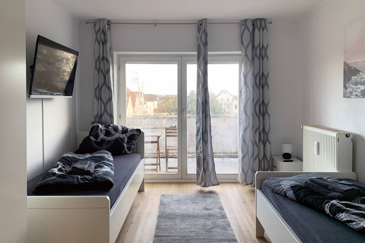A modern bedroom with a sofa bed, a single bed, a TV mounted on the wall, and a view of the outside through large windows.