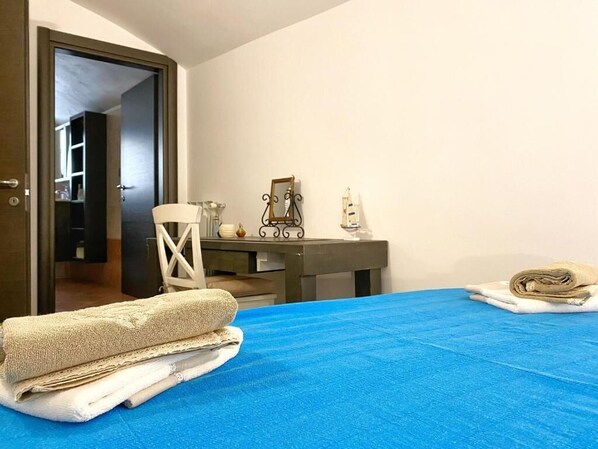 A bedroom with a blue bedspread, a chair, a desk with a mirror, and a door leading to another room. - Donna Lucrezia (Monopoli)