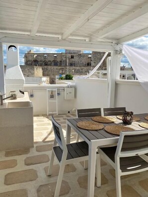An outdoor patio with a table set for dining, surrounded by white furniture and a stone floor. - Donna Lucrezia (Monopoli)